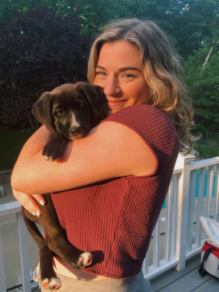 Jordan, long blonde hair with blue eyes and smiling, is holding her small black colored puppy in her arms. 