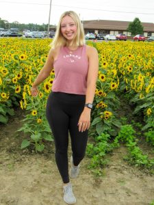 Allison is stadning in front of a field of sunflowers. She has medium length blonde hair, a pink shirt, and black yoga pants and is smiling at the camera. 