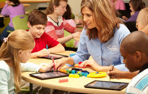 Three young kids and a female teacher sitting around a table. Kids are looking at their tablets lying on the table while teacher is smiling at kid in the center.