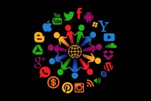 An image of two circles. The external circle in composed of different icons of social media and multimedia. The internal circle consist of people holding hands around a globe. 